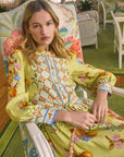 A person with long hair sits on a floral-patterned chair, wearing the Lillian Dress by Hunter Bell NYC—a green, button-front dress featuring intricate blue and orange fruit and geometric patterns. The cozy setting beautifully complements the floral-themed decor.