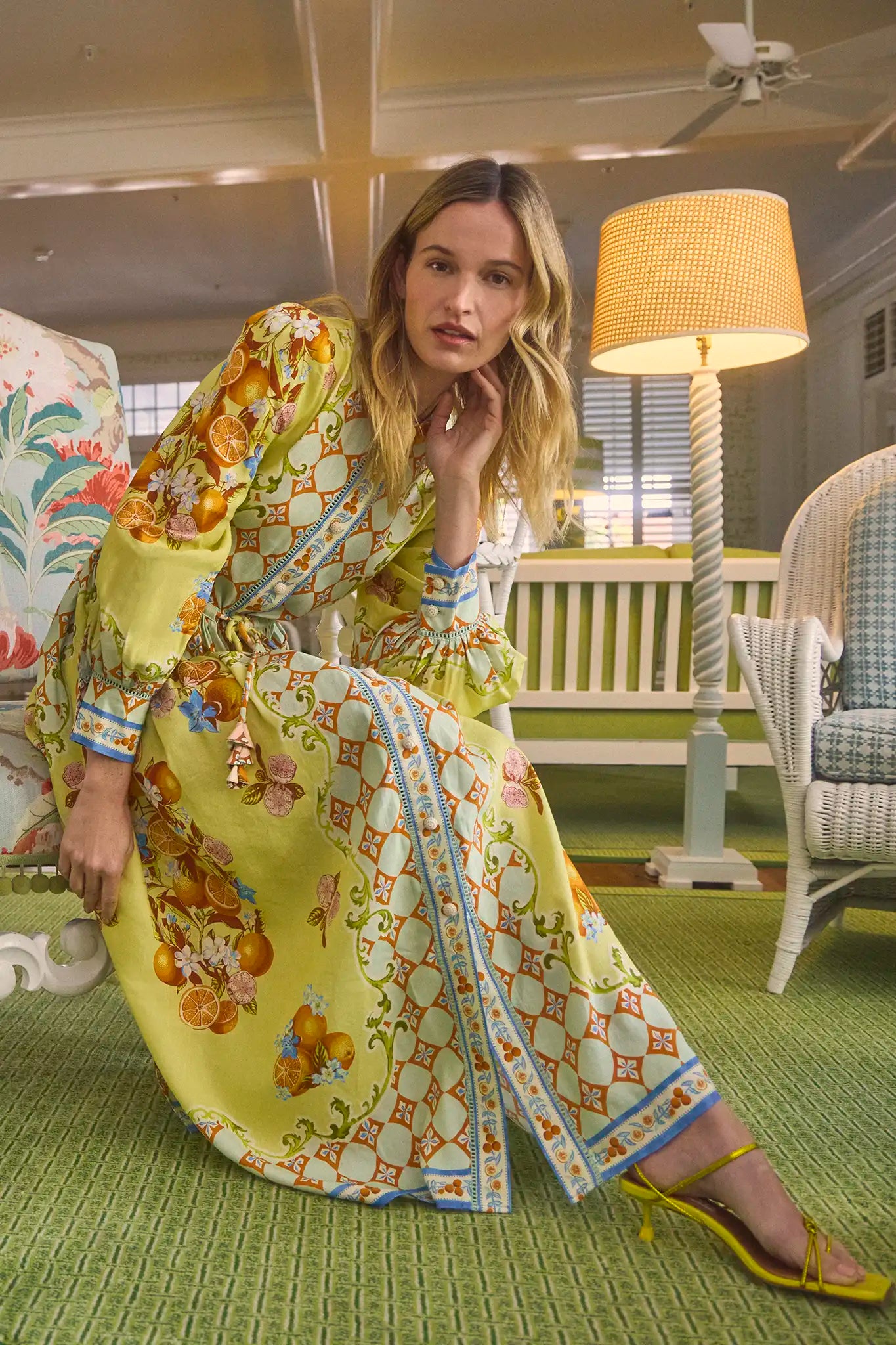 A woman in a Hunter Bell NYC Lillian Dress, a long button-front yellow dress with a drawstring waist, poses indoors on a cushioned bench. The room's green carpet, floral patterns, wicker chair, and tall lamp create a cozy vintage ambiance.