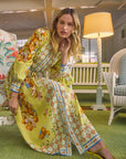 A woman in a Hunter Bell NYC Lillian Dress, a long button-front yellow dress with a drawstring waist, poses indoors on a cushioned bench. The room's green carpet, floral patterns, wicker chair, and tall lamp create a cozy vintage ambiance.