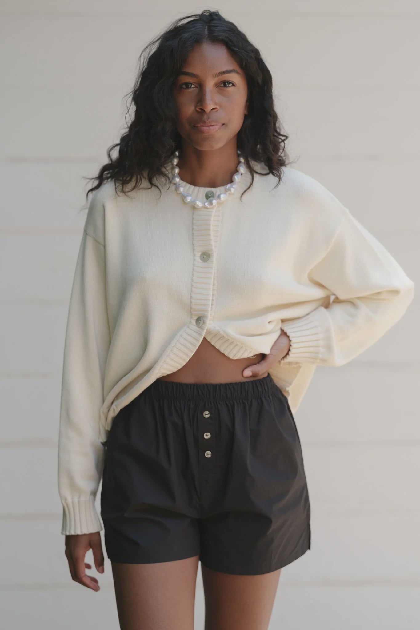 A person with wavy hair is wearing a pearl necklace, The Pop Boxer by Donni in black, and a cream-colored cotton poplin cardigan with a relaxed fit. They are against a neutral background, the cardigan is partially buttoned, and one hand rests in a pocket.