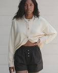 A person with wavy hair is wearing a pearl necklace, The Pop Boxer by Donni in black, and a cream-colored cotton poplin cardigan with a relaxed fit. They are against a neutral background, the cardigan is partially buttoned, and one hand rests in a pocket.