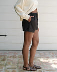 A person in a relaxed fit outfit featuring Donni's cream sweater, The Pop Boxer black shorts, and brown sandals stands on a brick surface. Perfect for a sunny day, the attire suggests breathable comfort. Only the lower half is visible.