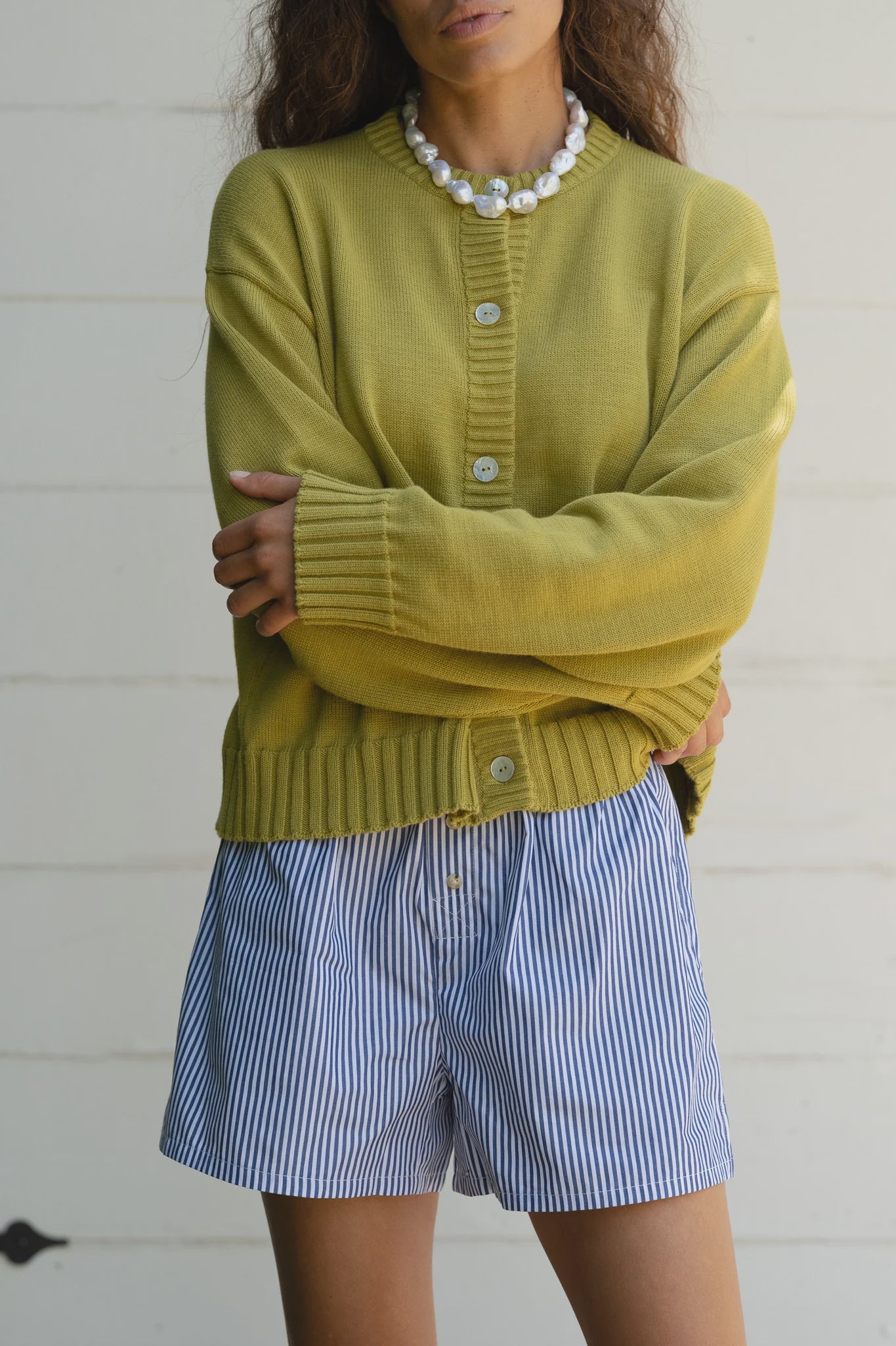 A person in a relaxed-fit yellow cardigan and Donni's The Stripe Pop Boxer shorts crosses their arms. Their long, wavy hair complements a pearl necklace against a plain, light-colored wall, showcasing an effortless Los Angeles style.