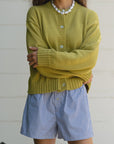 A person in a relaxed-fit yellow cardigan and Donni's The Stripe Pop Boxer shorts crosses their arms. Their long, wavy hair complements a pearl necklace against a plain, light-colored wall, showcasing an effortless Los Angeles style.