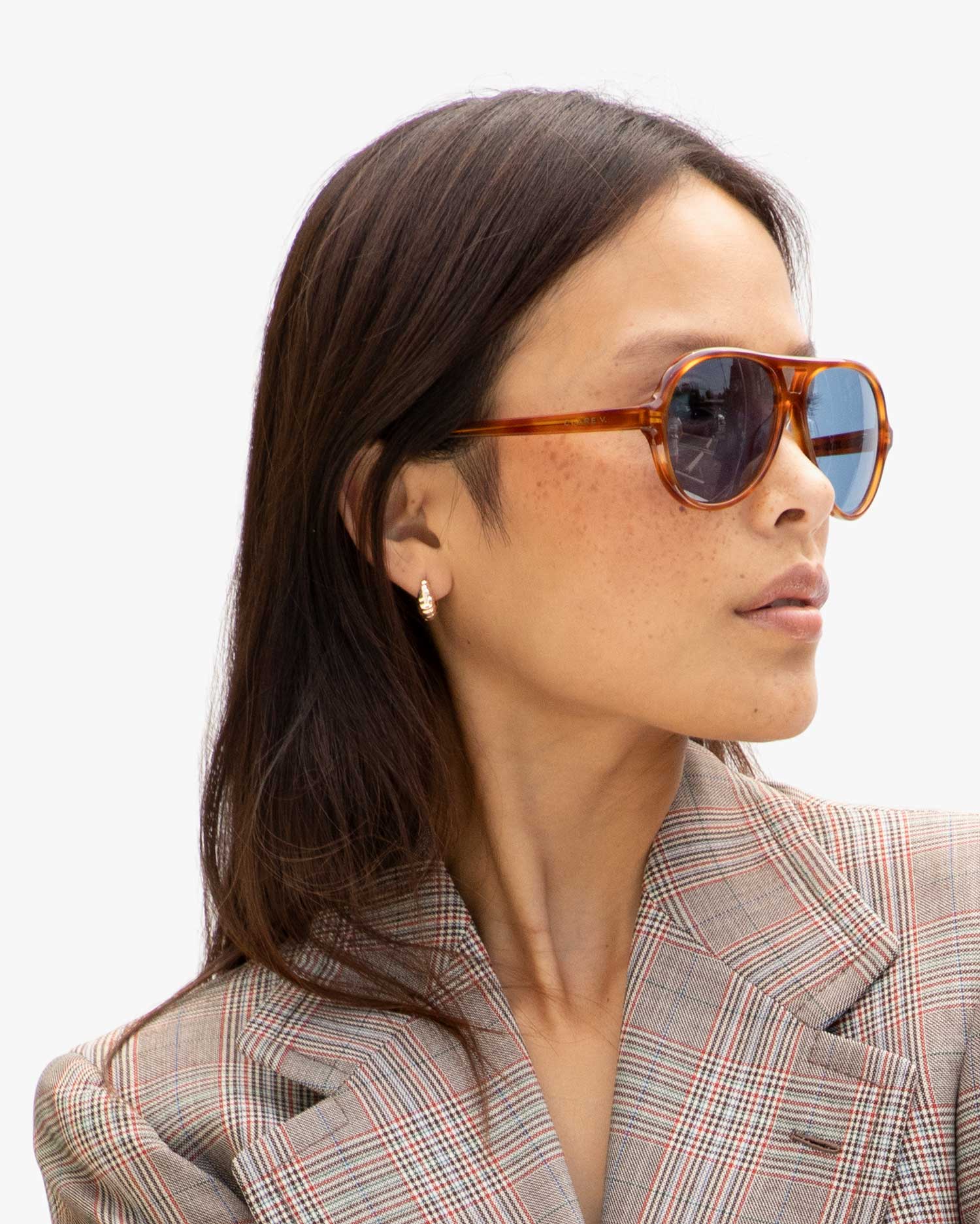 A person with long dark hair wearing Clare Vivier&#39;s eco-friendly Lizzie Sunglasses in tortoiseshell and a plaid blazer looks to the side against a plain background. The sunglasses, crafted from recycled bio-based materials, offer stylish UVA/UVB protection as they wear small hoop earrings.