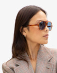 A person with long dark hair wearing Clare Vivier's eco-friendly Lizzie Sunglasses in tortoiseshell and a plaid blazer looks to the side against a plain background. The sunglasses, crafted from recycled bio-based materials, offer stylish UVA/UVB protection as they wear small hoop earrings.