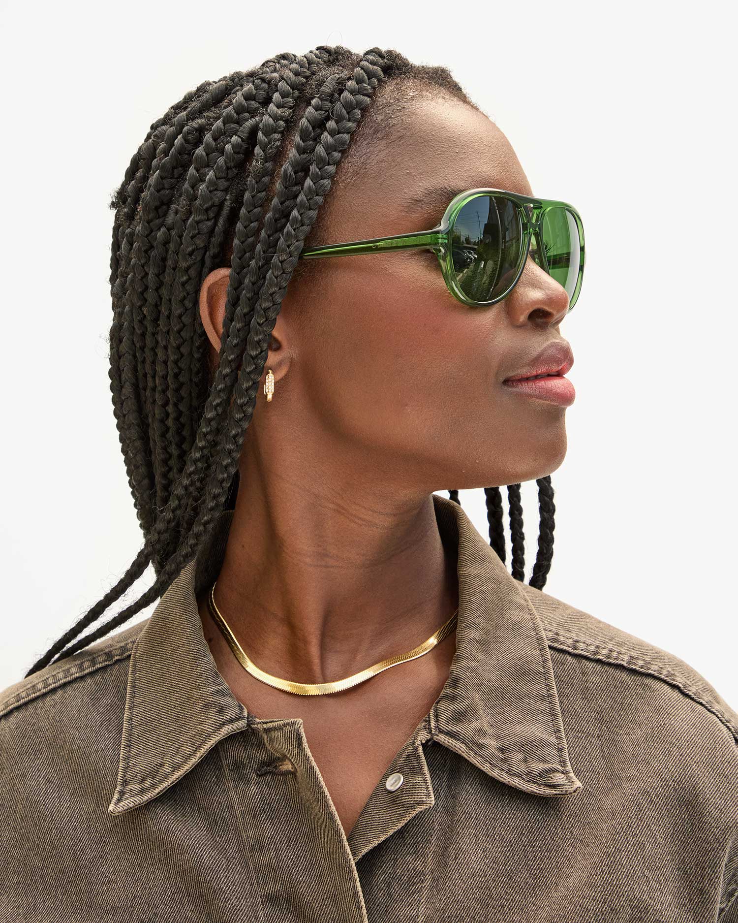 A person wearing Clare Vivier&#39;s Lizzie Sunglasses, which offer UVA/UVB protection, and a brown jacket with braided hair looks to the side. They are accessorized with a gold necklace and hoop earrings. The background is blurred and light gray, emphasizing their stylish commitment to sustainability.