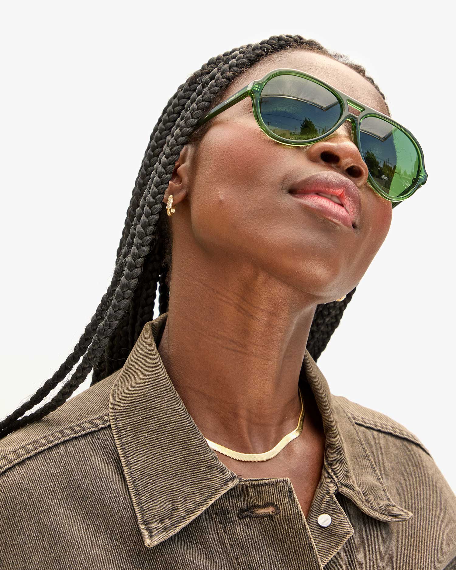 Wearing braided hair, a person dons the Lizzie Sunglasses by Clare Vivier, made from recycled bio-based materials in a vibrant green shade and pairs them with a brown denim jacket, while gazing upwards. A gold necklace and small hoop earrings add a subtle sparkle against the plain, light-colored background.