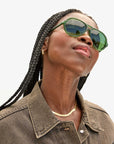 Wearing braided hair, a person dons the Lizzie Sunglasses by Clare Vivier, made from recycled bio-based materials in a vibrant green shade and pairs them with a brown denim jacket, while gazing upwards. A gold necklace and small hoop earrings add a subtle sparkle against the plain, light-colored background.