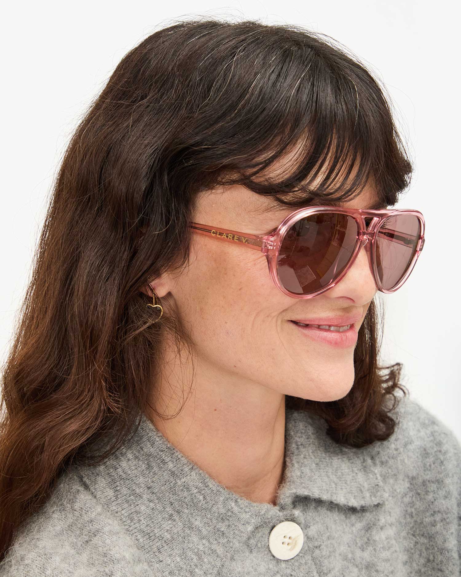 A brown-haired woman with a light complexion smiles in Lizzie Sunglasses by Clare Vivier, made from recycled materials. She wears a gray coat and small hoop earrings against a plain white background.