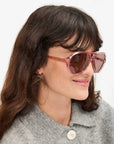 A brown-haired woman with a light complexion smiles in Lizzie Sunglasses by Clare Vivier, made from recycled materials. She wears a gray coat and small hoop earrings against a plain white background.