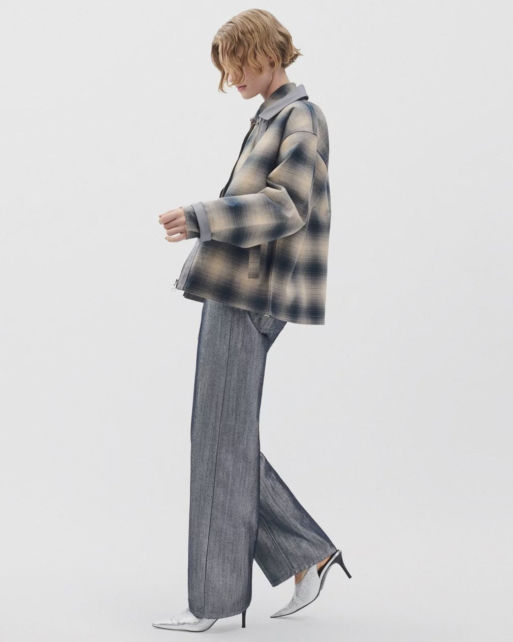 A person with short wavy hair stands sideways against a white background in an oversized plaid jacket, Rag & Bone logan wide -metallic indigo jeans, and pointed white heels.