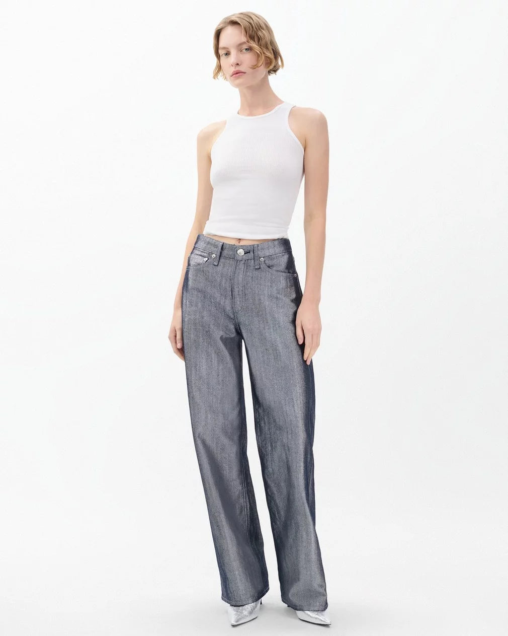 A woman with short blonde hair wears a white sleeveless tank top and Rag & Bone's Logan Wide jeans in metallic indigo, standing against a plain white background with a neutral expression.