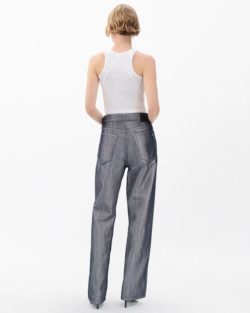 A person with short blonde hair models Rag & Bone's logan wide -metallic indigo jeans, styled with a white sleeveless top, standing with their back to the camera against a plain white background.