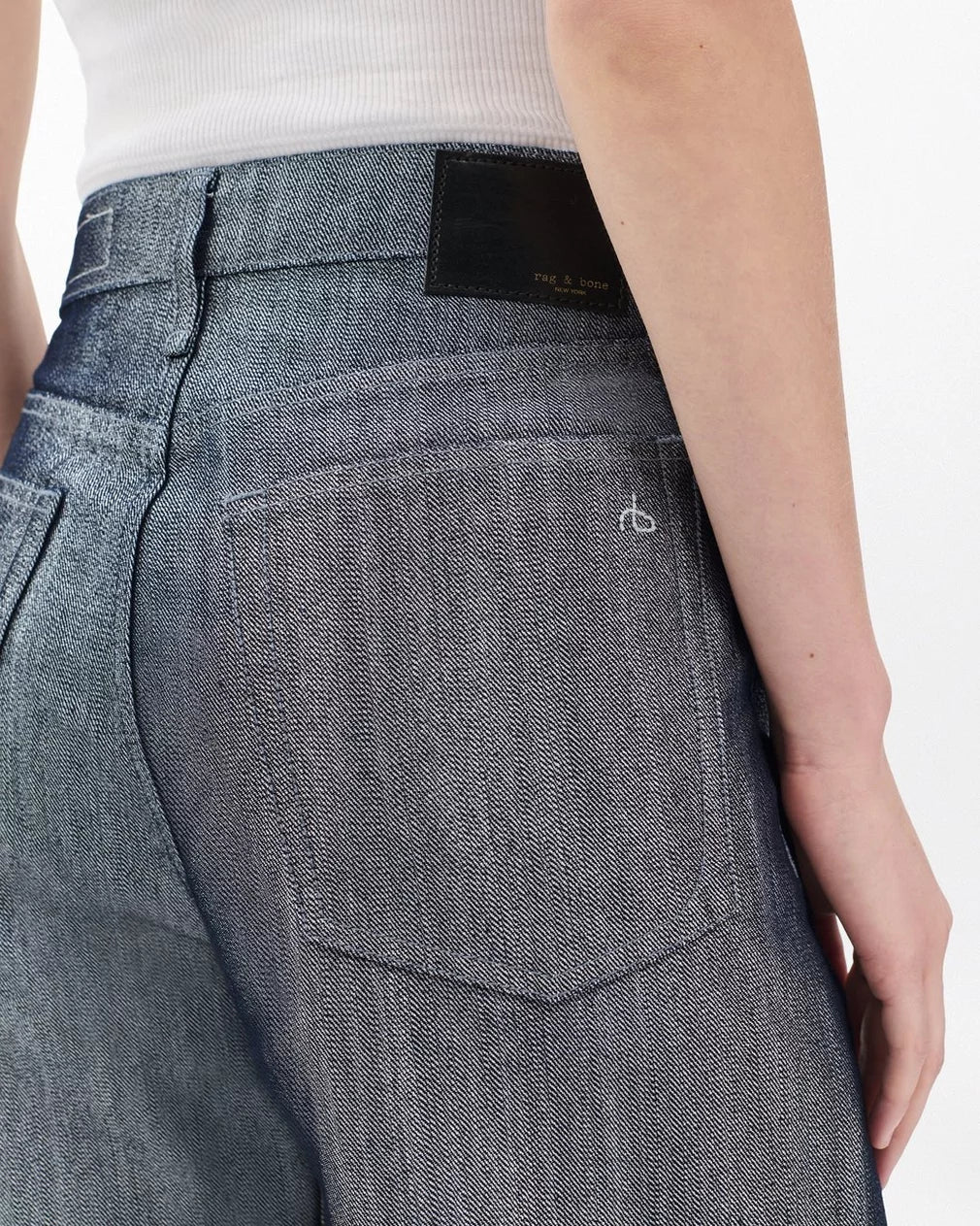 A person wears Rag & Bone's logan wide -metallic indigo jeans in lightweight, rigid denim with a faded gray-blue wash and a white ribbed tank top. The image highlights the jeans’ back pocket with a black waistband label and white stitching.