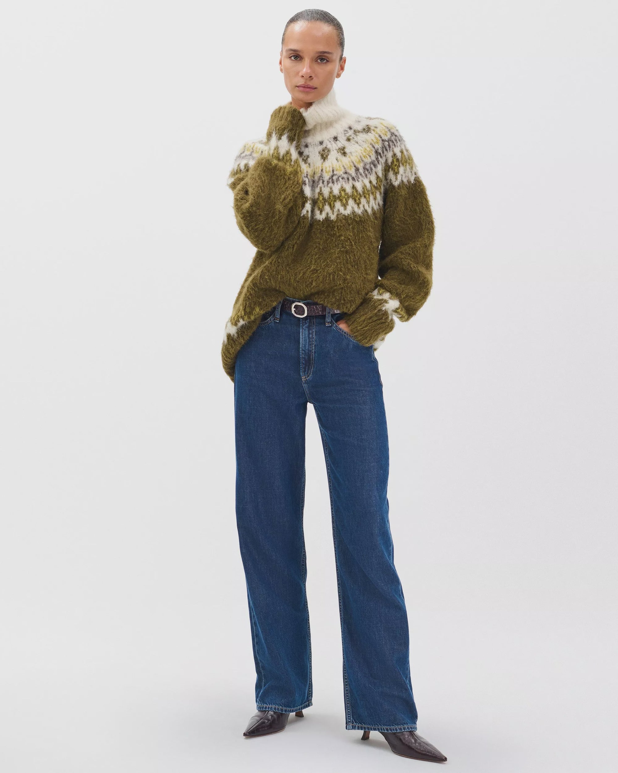 A woman stands against a white background wearing Rag & Bone ftwt logan mr wide leg jeans in dark indigo, paired with a fuzzy patterned sweater in olive green, white, and beige, and dark pointed-toe boots.