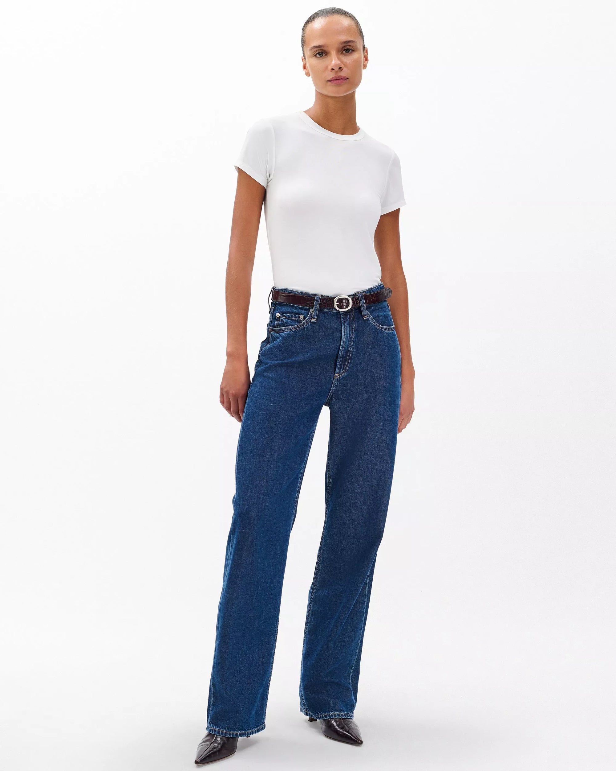 A woman stands against a white background wearing Rag & Bone's ftwt logan mr wide leg jeans, a fitted white short-sleeve t-shirt, a black belt, and black pointed-toe shoes.