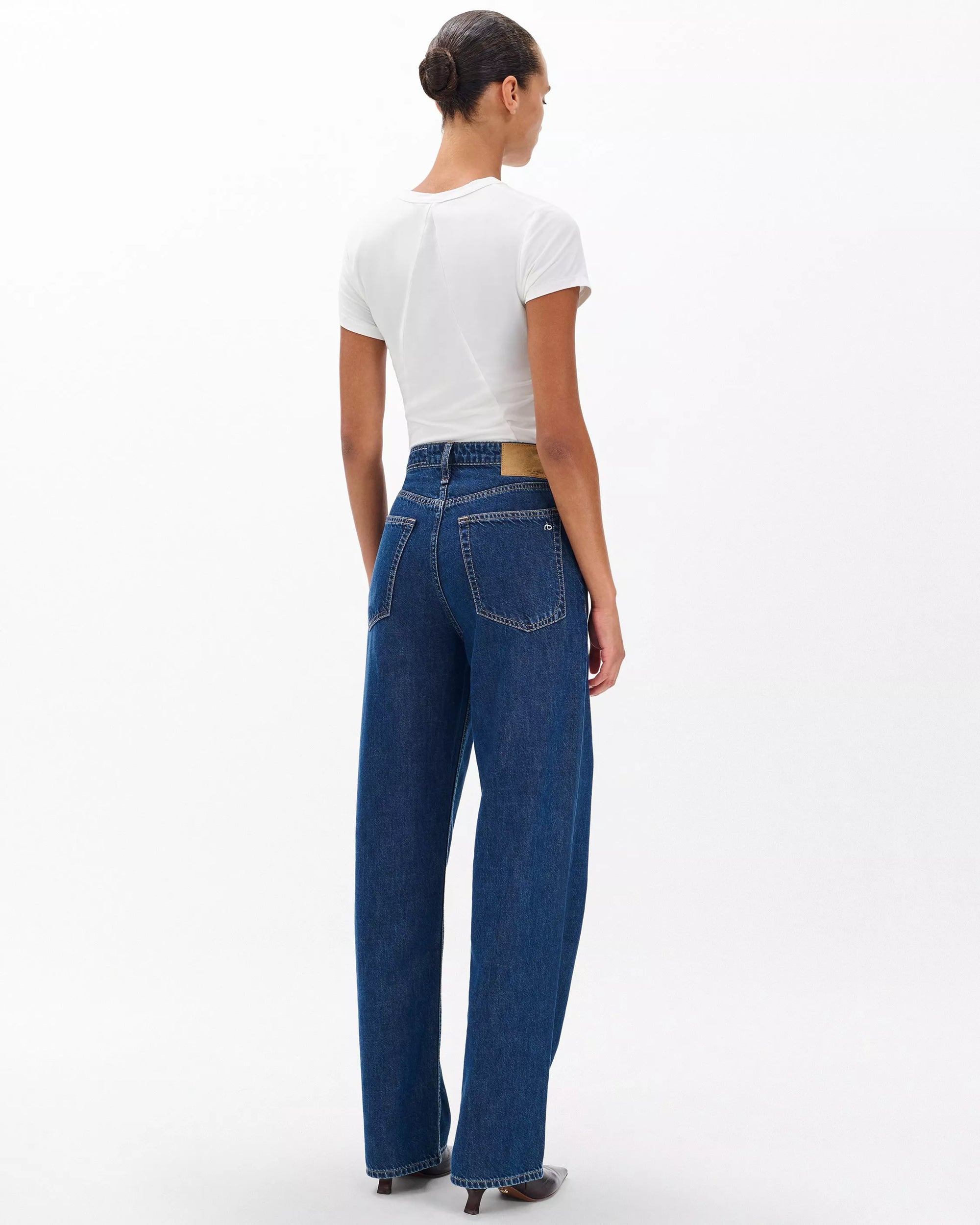 A person with dark hair in a bun faces away, wearing a fitted white tee and Rag & Bone ftwt logan mr wide leg jeans in dark indigo, standing against a plain white background.