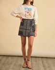 A woman stands on a wooden floor wearing the Hunter Bell NYC "I'm That Cowgirl LS Tee" tucked into a black bandana-print wrap skirt and black strappy sandals. Her chic, cowgirl-inspired style is showcased, though her face remains unseen.