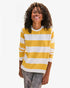 A curly-haired person smiles with closed eyes, wearing a Clare Vivier Long Sleeve Tee with wide yellow and white stripes described as reminiscent of sunny Portugal, paired with faded jeans against a plain white background.