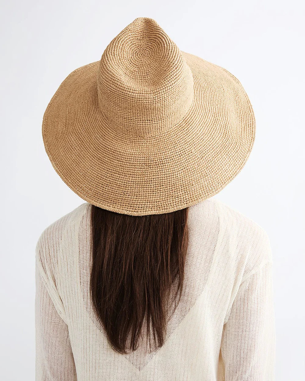 A person with long brown hair wears the Janessa Leone Waverly Sand raffia straw hat and a sheer light top, photographed from behind against a white background.