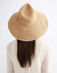 A person with long brown hair wears the Janessa Leone Waverly Sand raffia straw hat and a sheer light top, photographed from behind against a white background.