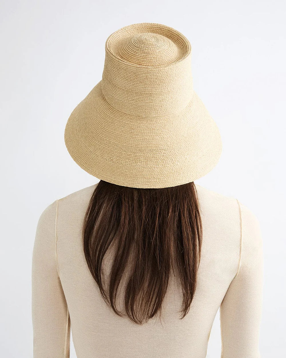 A person with long brown hair and a cream top is seen from behind, wearing the Janessa Leone Suni Natural handcrafted Madagascar hat against a plain light background.
