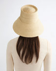 A person with long brown hair and a cream top is seen from behind, wearing the Janessa Leone Suni Natural handcrafted Madagascar hat against a plain light background.
