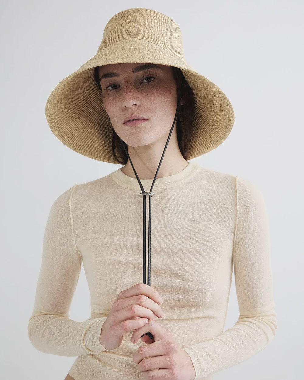 Wearing the Janessa Leone Suni Natural handcrafted Madagascar sun protection hat and a long-sleeved beige top, a person stands against a plain light background, holding the hat's black chin strap with both hands.