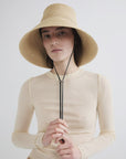Wearing the Janessa Leone Suni Natural handcrafted Madagascar sun protection hat and a long-sleeved beige top, a person stands against a plain light background, holding the hat's black chin strap with both hands.