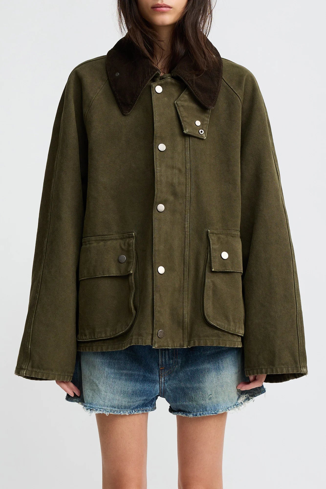 A woman wears the Denimist O/S Field Jacket in sturdy cotton—olive green with a dark brown corduroy collar, large front pockets, and silver snaps—paired with denim shorts. Her face is partially out of frame against a plain background.