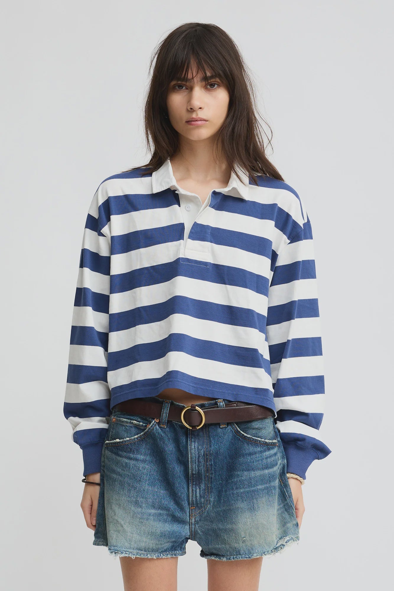 A young woman with long brown hair wears the Denimist Rugby LS Tee in Blue/Ecru Stripe, styled with denim shorts and a brown belt, standing against a plain light background.