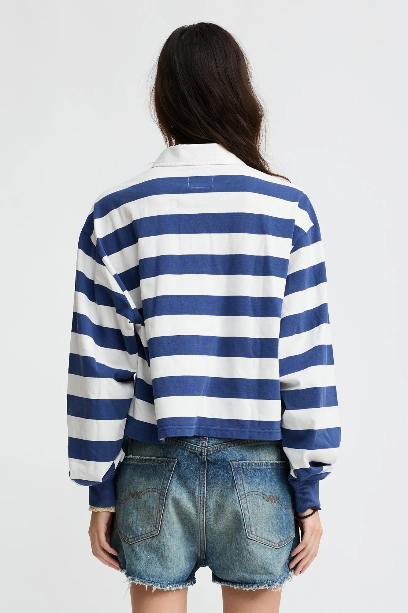 A woman with brown hair wears the Denimist Rugby LS Tee in blue ecru stripe and frayed denim shorts, facing away against a plain white background.