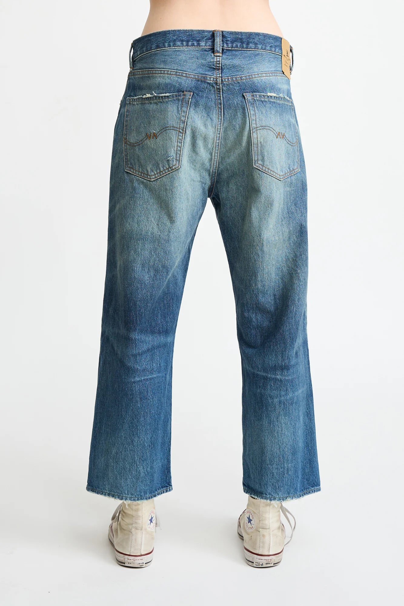 Back view of a person wearing Denimist HAISLEY SLOUCH BF JEAN in faded blue with wide legs and frayed hems, paired with white sneakers. Made from premium denim, shown against a plain white background.