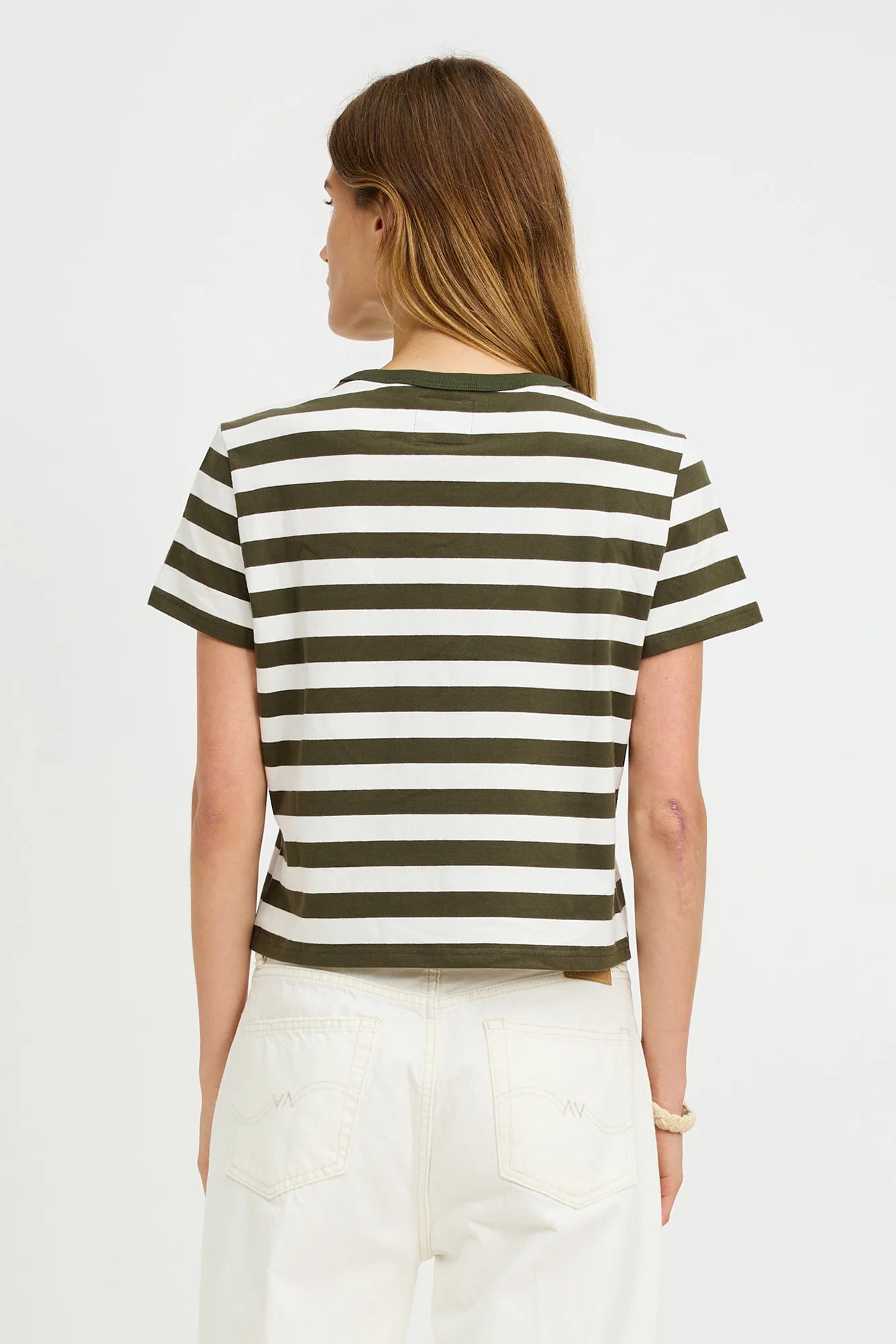 A woman with long brown hair faces away, wearing the Denimist STRIPED BABY T—a short-sleeve olive green and white crewneck tee—paired with high-waisted white jeans, set against a plain white background.