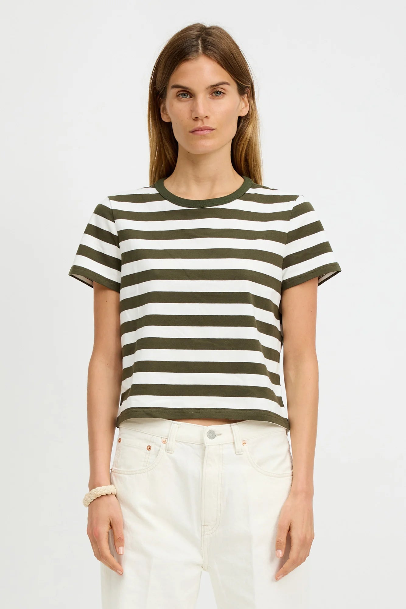 A woman with straight brown hair models the Denimist STRIPED BABY T, a short-sleeved black and white crewneck tee, paired with high-waisted white pants against a plain white background.