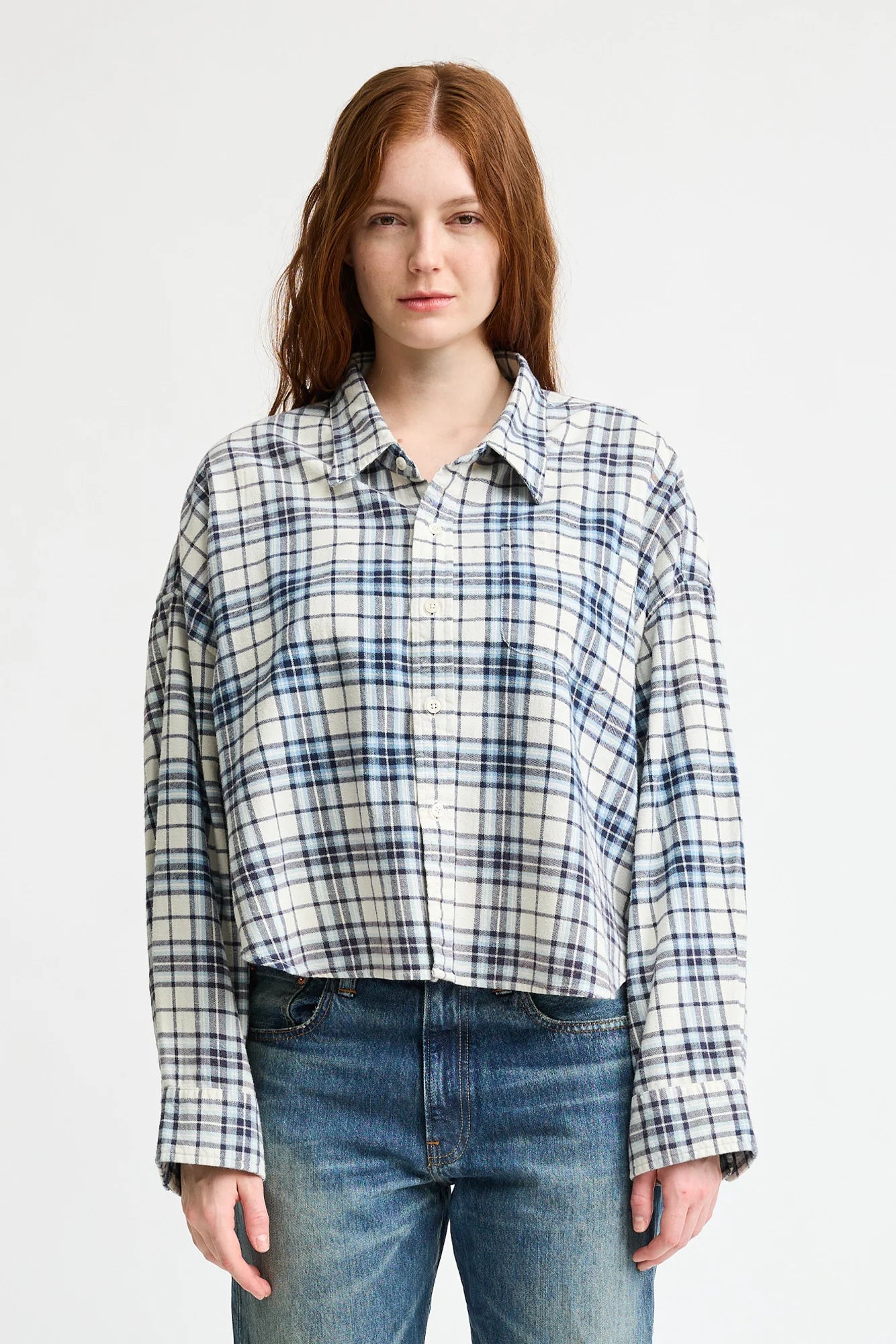 A person with long, reddish-brown hair wears the Denimist CROPPED BUTTON FRONT SHIRT in blue and white plaid with a boxy fit, paired with blue jeans, standing against a plain white background.