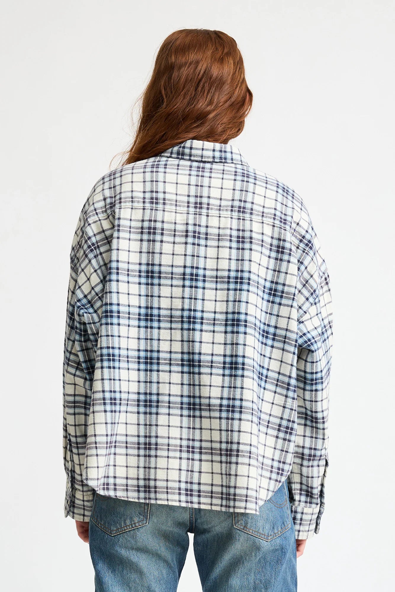 A person with long, wavy brown hair stands with their back to the camera against a plain light background, wearing Denimist's CROPPED BUTTON FRONT SHIRT and blue jeans.