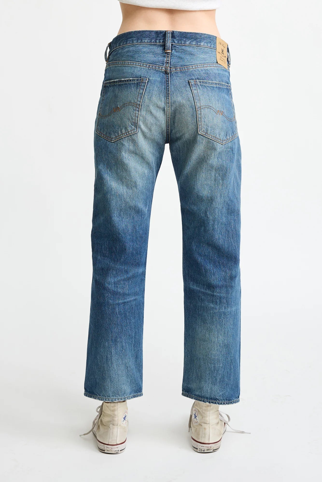 A person in Denimist LUCY BF JEAN ESHER BLUE, relaxed fit, and white sneakers stands facing away, showcasing the jeans’ two back pockets and leather patch on the waistband.