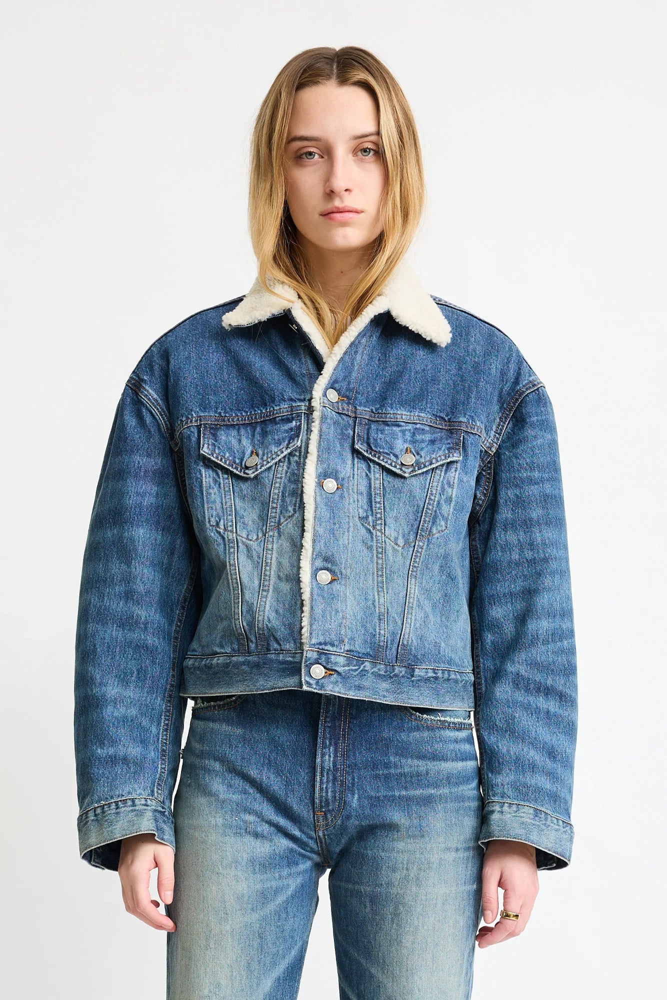 A person wearing the Denimist IRIS BOXY TRUCKER JACKET in indigo denim with a white shearling collar and blue jeans stands facing forward against a plain white background.