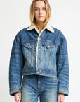 A person wearing the Denimist IRIS BOXY TRUCKER JACKET in indigo denim with a white shearling collar and blue jeans stands facing forward against a plain white background.