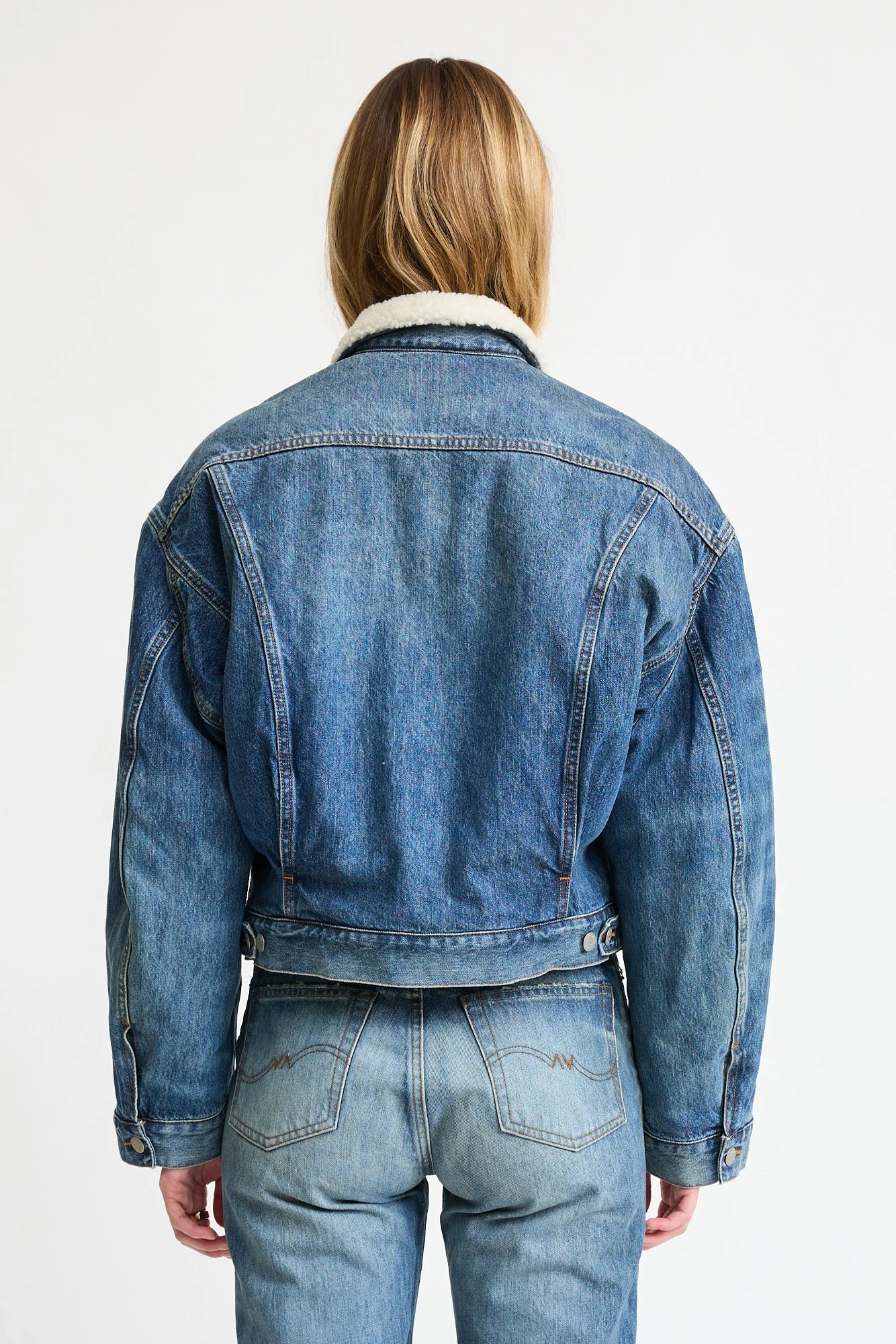 A person with light brown hair, wearing Denimist's IRIS BOXY TRUCKER JACKET in blue denim with a white fleece collar and matching jeans, stands with their back to the camera against a plain white background.