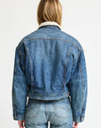 A person with light brown hair, wearing Denimist's IRIS BOXY TRUCKER JACKET in blue denim with a white fleece collar and matching jeans, stands with their back to the camera against a plain white background.