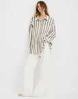 A person stands against a white background wearing the Denimist BUTTON FRONT SHIRT STRIPE, a relaxed fit, long-sleeve button-down with bold beige and white jumbo stripes, paired with wide-legged white pants.