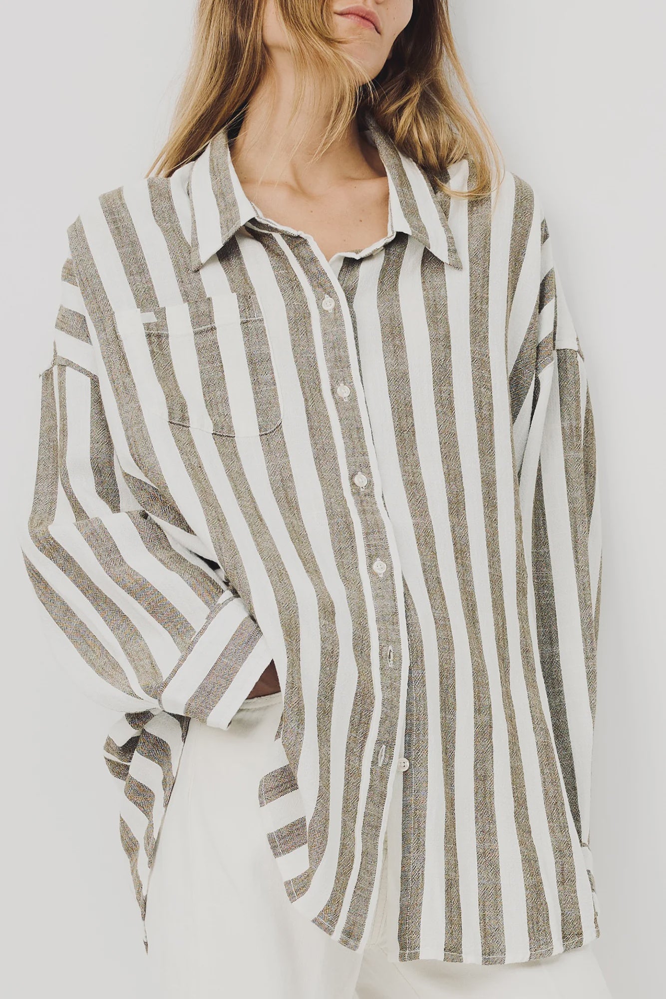 A woman models the Denimist BUTTON FRONT SHIRT STRIPE, a relaxed fit with jumbo beige and white stripes, paired with white pants. Her face is partially out of frame as she stands against a plain background.