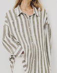 A woman models the Denimist BUTTON FRONT SHIRT STRIPE, a relaxed fit with jumbo beige and white stripes, paired with white pants. Her face is partially out of frame as she stands against a plain background.