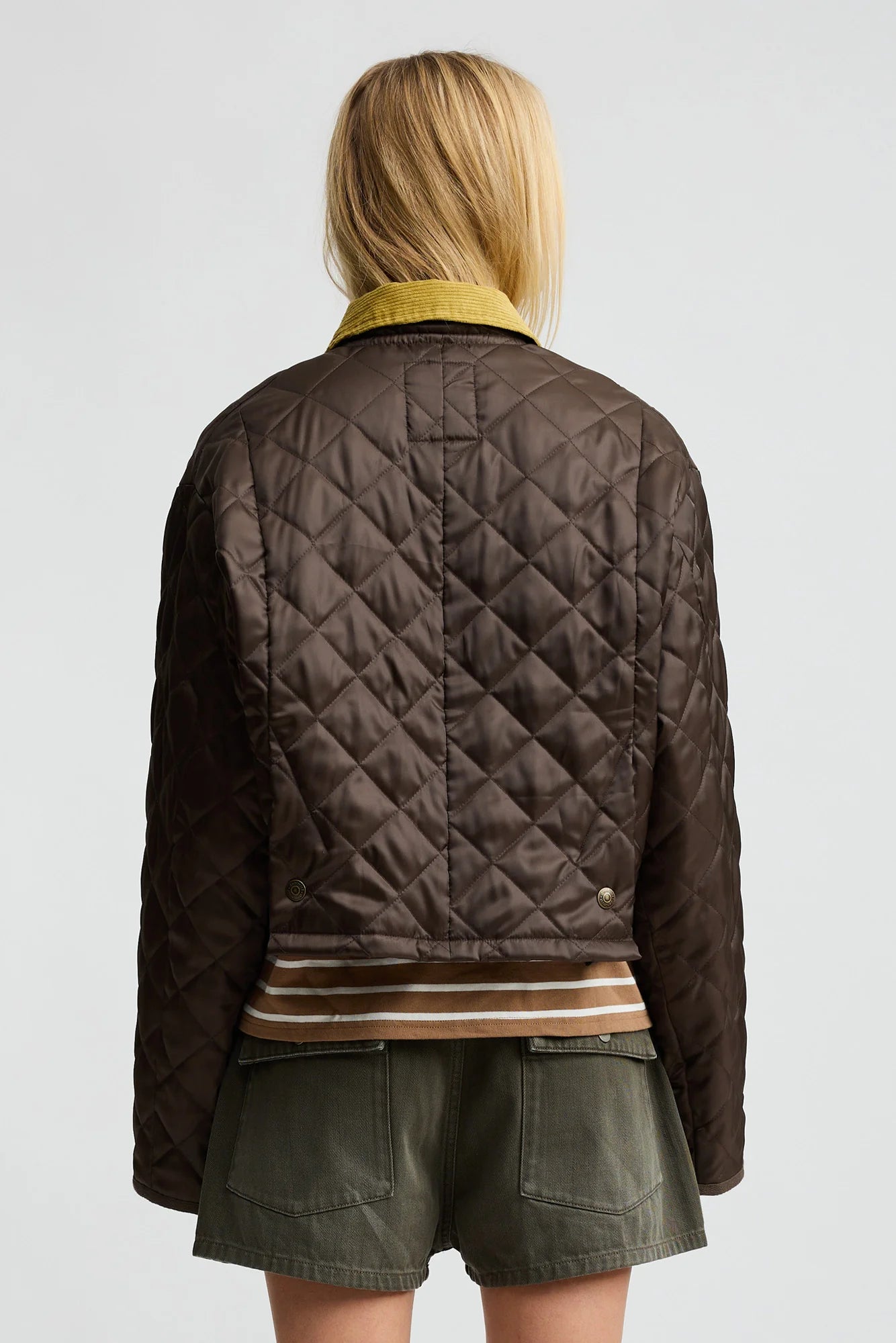 A person with blonde hair, seen from behind, wears the Denimist Cropped Quilted Jacket with a corduroy collar, a striped shirt, and olive green shorts against a plain light background.