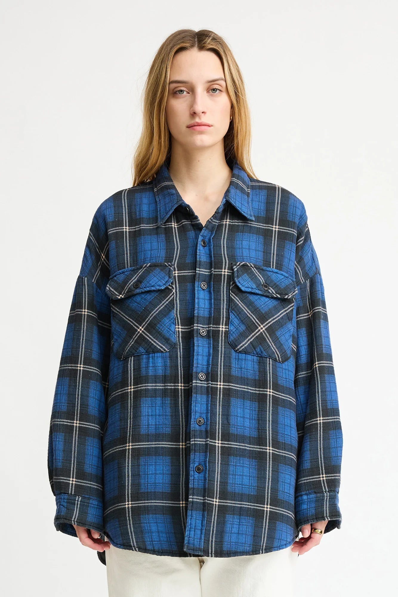 A person with long blonde hair wears the Denimist CPO SHIRT JACKET in an oversized blue and black plaid cotton style, paired with off-white pants, standing against a plain white background.
