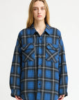 A person with long blonde hair wears the Denimist CPO SHIRT JACKET in an oversized blue and black plaid cotton style, paired with off-white pants, standing against a plain white background.