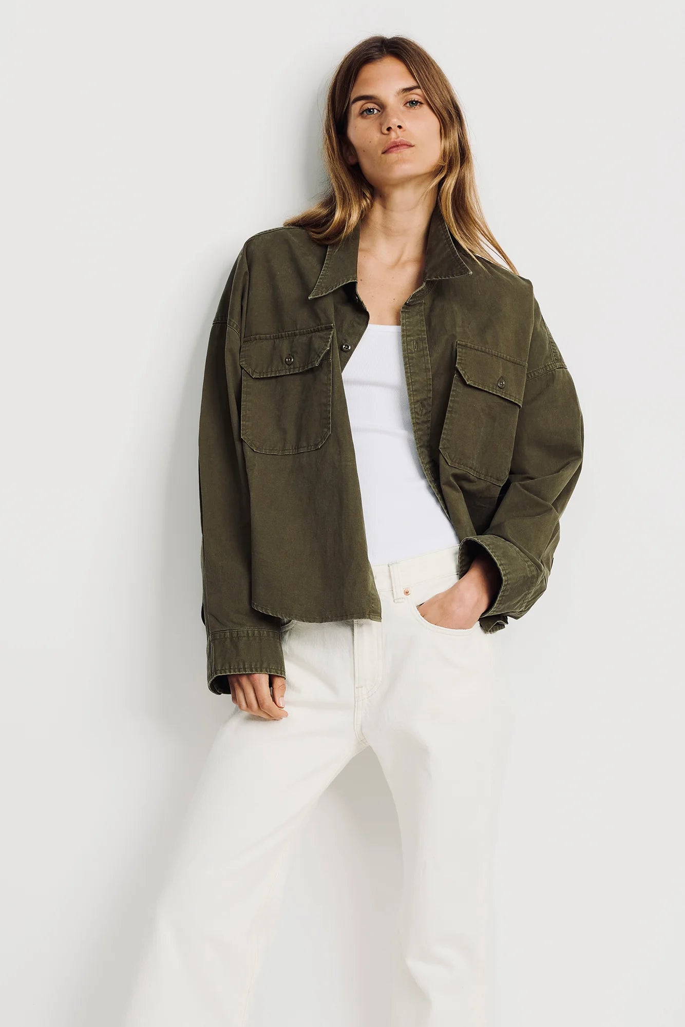 A woman poses against a white background wearing Denimist’s CROPPED UTILITY SHIRT in olive, styled with a white tank top and high-waisted white pants, one hand in her pocket and a relaxed expression.